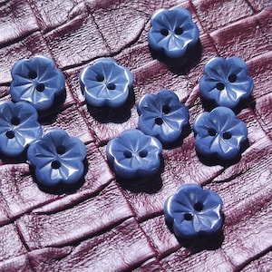 Puede incluir: Un conjunto de nueve botones pequeños de color azul con forma de flor. Cada botón presenta un diseño de cinco pétalos con un acabado liso y brillante, además de dos orificios centrales para su fijación. Los botones están dispuestos dispersos.