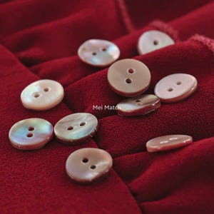 May include: A collection of iridescent, two-hole buttons in various sizes and shades of cream, brown, and iridescent colors. The buttons are scattered on a deep red fabric, creating a visually appealing contrast. The text "Mei Match" is visible.