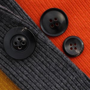 Puede incluir: Primer plano de tres botones negros con cuatro agujeros cada uno, dispuestos sobre una tela texturizada gris, naranja y amarilla. Los botones varían en tamaño, con el botón más grande a la izquierda.