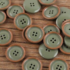 Op de afbeelding: Een verzameling olijfgroene knopen met elk vier gaten en een bruine rand. De knopen zijn gerangschikt op een houten oppervlak, wat een textuurrijke en aantrekkelijke opstelling creëert.