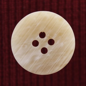 May include: A white, round button with four holes in a cross pattern. The button has a slightly textured surface.