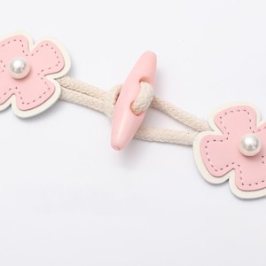 Pode incluir: Dois botões rosa em forma de flor com centros brancos e detalhes de pérola conectados por um cordão branco e um botão de alternância de plástico rosa.