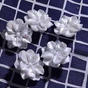 May include: Five white, flower-shaped decorative embellishments. Each flower has multiple petals and a textured surface, creating a realistic appearance. The flowers are arranged on a dark blue fabric with a white grid pattern.