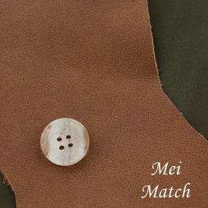 Puede incluir: Un botón blanco con cuatro agujeros, sobre un fondo de cuero marrón. El texto "Mei Match" es visible en la esquina inferior derecha de la imagen.