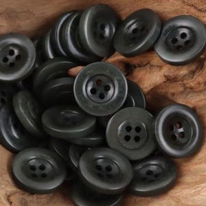 May include: A collection of round, dark gray buttons, each featuring four central holes for attachment. The buttons have a smooth, slightly reflective surface and are arranged in a dense, overlapping pile, showcasing their uniform circular shape.