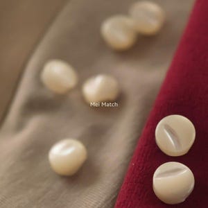 May include: A collection of small, round, off-white buttons with a smooth, pearlescent finish. Some buttons are scattered, while two are placed side-by-side on a red fabric edge. The text Mei Match is printed in the center of the image.