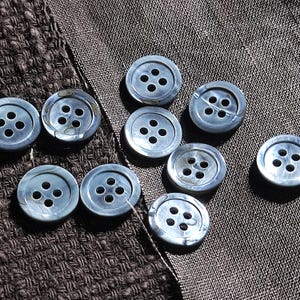 May include: A collection of ten round, four-hole buttons with a smooth, iridescent gray finish. Each button features a slightly recessed center and a raised outer rim, displaying a subtle pearlescent sheen across the surface of the material.