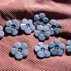 Puede incluir: Una colección de diez botones con forma de flor en un color gris azulado apagado. Cada botón presenta cinco pétalos redondeados y cuatro orificios centrales para coser. Los botones tienen una textura superficial lisa y ligeramente veteada.