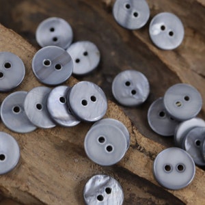 Peut inclure: Un ensemble de boutons ronds gris à deux trous avec une finition nacrée. Les boutons sont éparpillés sur une surface en bois.