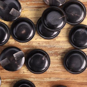 以下が含まれることがあります： 光沢のある黒い丸いコースターの俯瞰図。一部のコースターには、長方形のテクスチャ加工されたディテールがあります。 コースターは明るい茶色の木製の表面に配置されており、素朴な美学を作り出しています。