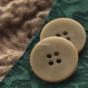 Op de afbeelding: Twee ronde, olijfkleurige knopen met een gladde afwerking. De voorste knoop heeft vier gaten, terwijl de gedeeltelijk zichtbare knoop erachter drie gaten heeft. De tekst "Mei Match" is in het wit op de voorste knoop gedrukt.