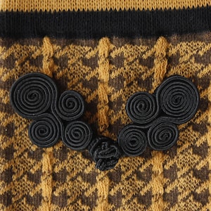 Könnte beinhalten: Nahaufnahme eines Kleidungsstücks mit einem Hahnentrittmuster in Braun- und Goldtönen. Das Kleidungsstück hat schwarze Verzierungen und dekorative schwarze Spiralverschlüsse. Der Stoff scheint gestrickt oder gewebt zu sein.