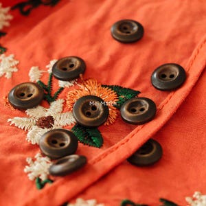 May include: Close-up of orange fabric with embroidered floral details and dark brown buttons. The buttons are round with two holes and are scattered across the fabric. The fabric has a textured appearance.