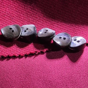 Puede incluir: Cinco botones redondos con un acabado brillante jaspeado en gris oscuro y negro. Cada botón presenta una superficie ligeramente abombada y dos orificios centrados para su fijación. Los botones están dispuestos en una fila horizontal.