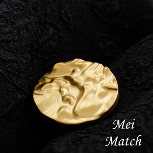 May include: A gold-toned brooch with a textured, abstract design. The brooch is shaped like a circle and has a raised, three-dimensional design.