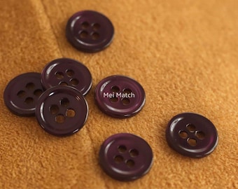May include: A collection of six dark purple buttons with four holes each, scattered on a tan fabric surface. The buttons have a glossy finish and are evenly spaced. One button has the text "Mei Match".