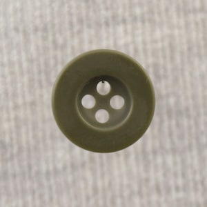 Op de afbeelding: Close-up van een ronde, olijfgroene knoop met vier gaatjes. De knoop heeft een licht verhoogde buitenrand en staat tegen een zachte, getextureerde, lichtgrijze achtergrond. Geschikt voor naaiprojecten.