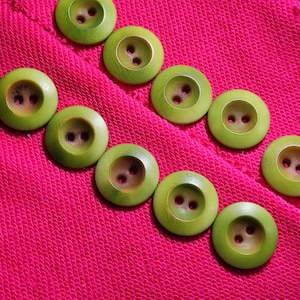 May include: A series of round, light green buttons with two central holes arranged in a diagonal line across a vibrant, textured hot pink fabric. Each button features a smooth, slightly concave surface with a subtle wood grain texture.
