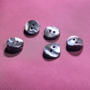 May include: Five circular buttons with a dark, iridescent, charcoal-grey finish and a smooth, slightly domed surface. Each button features two small, centered holes for attachment, displaying a subtle, swirling, pearlescent texture throughout.
