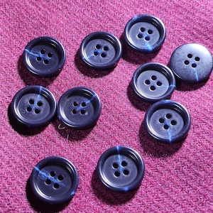 May include: Nine navy blue buttons with four holes each, arranged on a textured, deep pink fabric. Each button has a subtle blue accent line. The buttons are round and have a glossy finish.