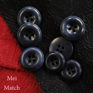 May include: A collection of seven dark blue plastic buttons with four holes each. The buttons are arranged in a semi-circle on a black fabric background.