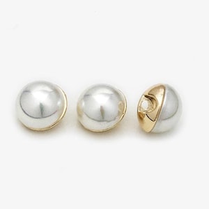 Może przedstawiać: Trzy półperłowe koraliki ze złotym akcentem. Koraliki są błyszczące, perłowo-białe ze złotą opaską na krawędzi. Jeden koralik ma złotą sekcję z otworem. Koraliki te nadają się do tworzenia biżuterii.