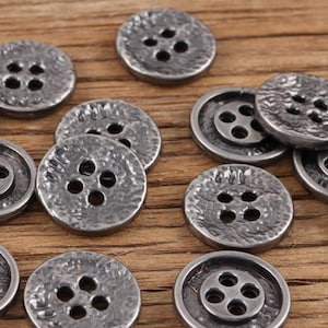 May include: A selection of antique silver-coloured buttons, each with four holes. The buttons have a textured, hammered finish and are displayed on a wooden surface. These buttons are suitable for sewing and craft projects.