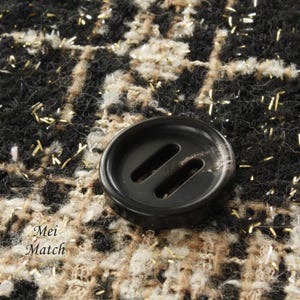May include: A black button with two parallel slits, set against a background of black, brown, and white tweed fabric with gold glitter accents.