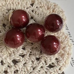 Puede incluir: Cinco pendientes esféricos de color burdeos con un acabado brillante. Cada pendiente tiene una base dorada. Los pendientes están colocados sobre un tapete de ganchillo color crema.