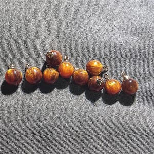 Könnte beinhalten: Eine Ansammlung von neun kugelförmigen Anhängern, die jeweils eine tief bernsteinfarbene Basis mit wirbelnden goldfarbenen Glitzereinschlüssen aufweisen. Jeder Anhänger ist mit einer kleinen, metallisch silberfarbenen Öse zur Befestigung versehen.
