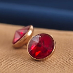 May include: A pair of round stud earrings featuring faceted, deep red stones set in polished gold-tone metal frames. The text Mei Match is visible on the surface of the front earring. The design is circular with a raised, multi-faceted centre.