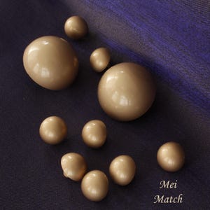 May include: A selection of beige, glossy pearls of different sizes arranged on a dark blue fabric. The pearls are smooth and spherical. The text "Mei Match" is visible in the bottom right corner.