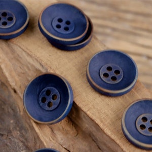 Può includere: Primo piano di diversi bottoni blu scuro con bordo dorato. I bottoni sono disposti su una superficie di legno.