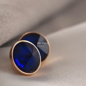 Puede incluir: Dos pendientes redondos de tipo botón con cristales facetados de color azul intenso engastados en monturas de metal de oro rosa pulido. El texto Mei Match es visible entre los dos pendientes circulares, que tienen una superficie lisa y brillante.