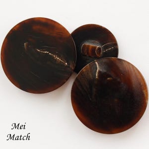 Puede incluir: Tres botones redondos con un patrón jaspeado de color marrón oscuro similar al carey y un acabado liso y brillante. Un botón está colocado para mostrar el vástago en la parte posterior. El texto "Mei Match" está impreso en negro abajo a la izquierda.