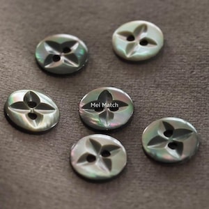 Puede incluir: Seis botones redondos de nácar iridiscente con un diseño de cuatro agujeros y un patrón de superficie tallado en forma de pétalos. Los botones muestran una paleta de colores gris y verde oscuro brillante. El botón central incluye el texto Mei Match.