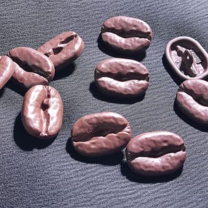 Puede incluir: Una colección de objetos marrones con forma de grano de café. Los objetos están dispuestos sobre una superficie texturizada de color gris oscuro. Los granos tienen un aspecto detallado y realista, con surcos y crestas.