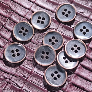 May include: Ten round, grey buttons with four holes each, displayed on a textured, burgundy surface. The buttons have a metallic sheen and a copper-coloured rim, creating a vintage aesthetic.