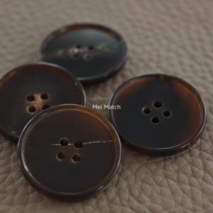 May include: A collection of four large, round buttons with a dark brown, tortoiseshell-like appearance. Each button has four holes for sewing. The buttons are arranged on a neutral-toned surface.