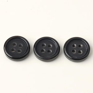 Puede incluir: Tres botones redondos negros con cuatro agujeros cada uno, dispuestos horizontalmente sobre un fondo blanco. Los botones tienen un acabado brillante y un borde ligeramente elevado. Cada botón mide unos 2,5 cm de diámetro.