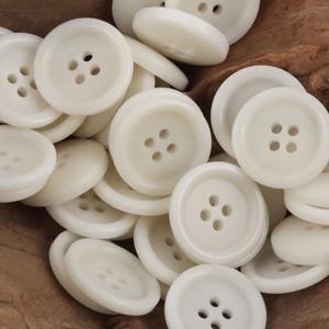 Może przedstawiać: Stosik białych plastikowych guzików, każdy z czterema otworami. Guziki są gładkie i mają lekko błyszczące wykończenie.