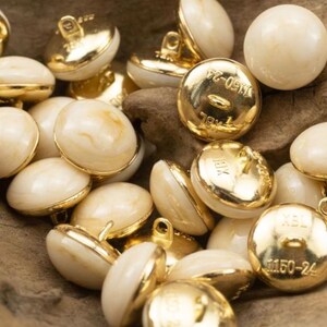 May include: A collection of white and gold buttons with a marbled effect. The buttons have a gold rim and a shank on the back. The buttons are marked with the text "M150-24" and "78x1".