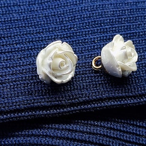 May include: Two white pearl-like rose buttons with gold accents. The buttons are on a blue knitted fabric.