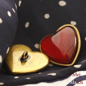 May include: Two heart-shaped buttons, one gold and one red. The gold button has a loop on top. The red button has a shiny, glass-like finish.