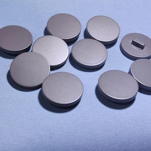 Puede incluir: Una colección de diez objetos metálicos grises circulares, posiblemente tapas o tapones, dispuestos sobre una superficie azul claro. Un objeto tiene una pequeña protuberancia rectangular. Los objetos están espaciados uniformemente.