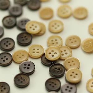 以下が含まれることがあります： 4つの穴の開いた茶色とベージュのボタンの詰め合わせ。ボタンは滑らかで光沢のある素材で作られています。