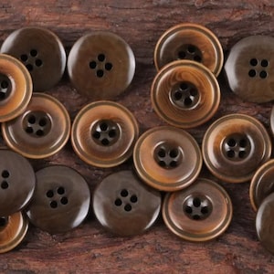 Peut inclure: Un assortiment de boutons marron, chacun percé de quatre trous. Les boutons varient en teinte, allant du marron clair au foncé, certains présentant un motif annulaire plus clair autour du centre. Parfaits pour la couture et les travaux manuels.