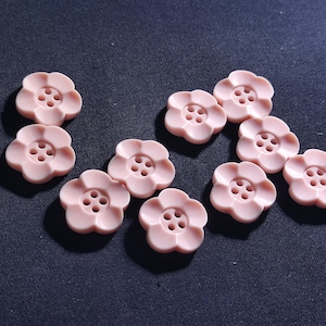Puede incluir: Un conjunto de botones rosas pálidos con forma de flor, con cuatro agujeros para coser. Están colocados sobre una tela azul oscuro. Estos botones decorativos son ideales para proyectos de costura y manualidades.