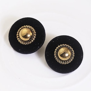 May include: Two black velvet stud earrings with a gold-colored center and a beaded border. The earrings are round and sit on a white surface. The earrings have a vintage or retro style.