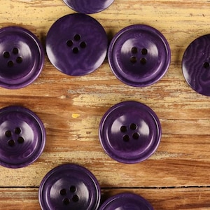 May include: Close-up of several large, round, purple buttons with four holes each. The buttons have a marbled appearance and are arranged on a weathered, light brown wooden surface. The buttons are a deep shade of purple.